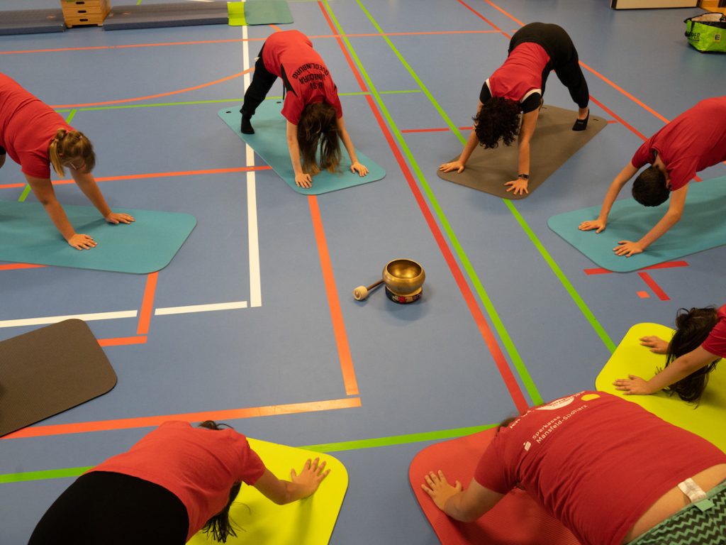 Die Matten liegen im Kreis. In der Mitte steht eine Klangschale. Die Teilnehmer zeigen die Yoga-Position "herabschauender Hund".